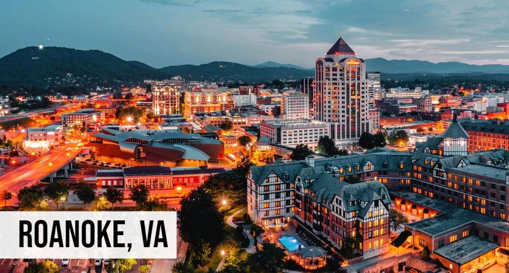Roanoke, Virginia nighttime cityscape with downtown buildings, streetlights, mountain backdrop, and surrounding valley landscape